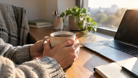 Hände halten eine Tasse Tee am Schreibtisch am Fenster am Morgen